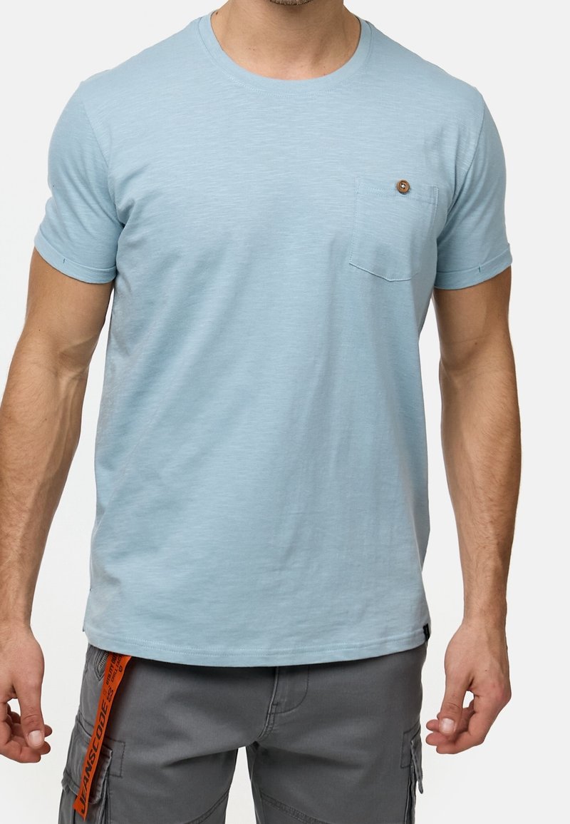 T-shirt bleu clair à manches courtes en tissu doux, avec un col rond et une poche à rabat ornementée d'un bouton décoratif.