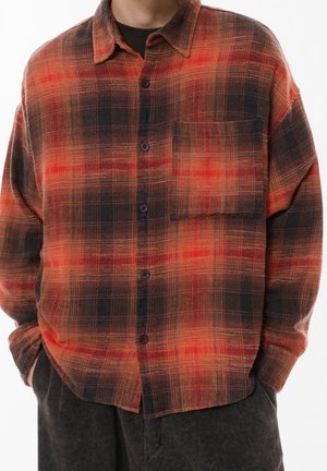 Homme portant une chemise ample à carreaux rouge et noir avec une poche poitrine et un pantalon foncé, les mains dans les poches, sur fond blanc.