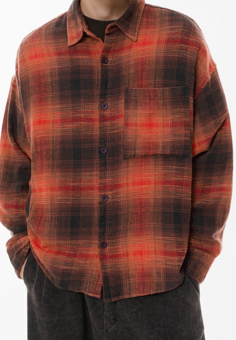 Homme portant une chemise ample à carreaux rouge et noir avec une poche poitrine et un pantalon foncé, les mains dans les poches, sur fond blanc.