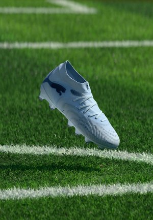 Chaussure de football blanche avec un logo de panthère noire en plein vol au-dessus d'une pelouse verte marquée de lignes blanches.