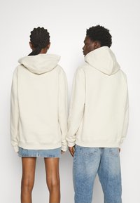 Dos personas llevan sudaderas con capucha beige, que presentan bolsillos tipo canguro y puños acanalados, combinadas con pantalones cortos de mezclilla y jeans.