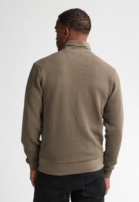 Kaki zip-up jacka med texturerad tyg, ribbade muddar och nederkant. Har en hög krage och bikakestruktur över hela kroppen.