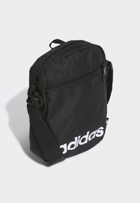 Bolso bandolera de tela negra con cierre de cremallera, que presenta un logo blanco de "adidas" y una correa ajustable. Diseño compacto y rectangular.