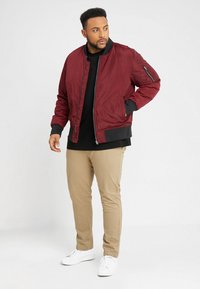 Giacca bomber bordeaux con polsini e orlo a coste, dotata di tasca con zip su manica, indossata sopra una maglietta nera, abbinata a pantaloni beigi e sneakers bianche.