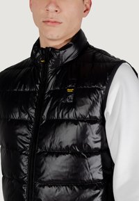 Chaleco acolchado negro con cuello alto, cierre de cremallera y textura acolchada. Logo destacado en el pecho, llevado sobre una camisa blanca.