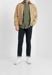 Tummanvihreän napitettavan paidan päällä on ruskea bomber-takki, tummansiniset farkut ja valkoiset tennarit. Takissa on joustavat hihansuut ja vetoketjusuljenta.