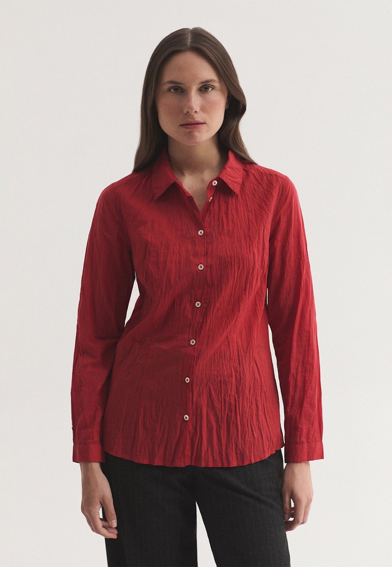 Femme aux longs cheveux bruns portant une chemise rouge à manches longues boutonnée et un pantalon noir, debout devant un fond clair uni.
