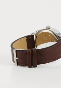 Bracelet de montre en cuir marron avec un boîtier en acier inoxydable argenté, équipé d'un cadran rond et d'une couture structurée le long des bords.