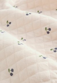 Tessuto trapuntato in morbido rosa con un motivo a diamante, caratterizzato da ciliegie blu ricamate e foglie verdi sparse qua e là.