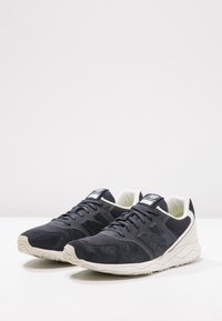 Námořnické modré sportovní boty s kombinací semiše a syntetických materiálů, s bílou podešví a texturovanými tkaničkami, navržené pro pohodlí a podporu.