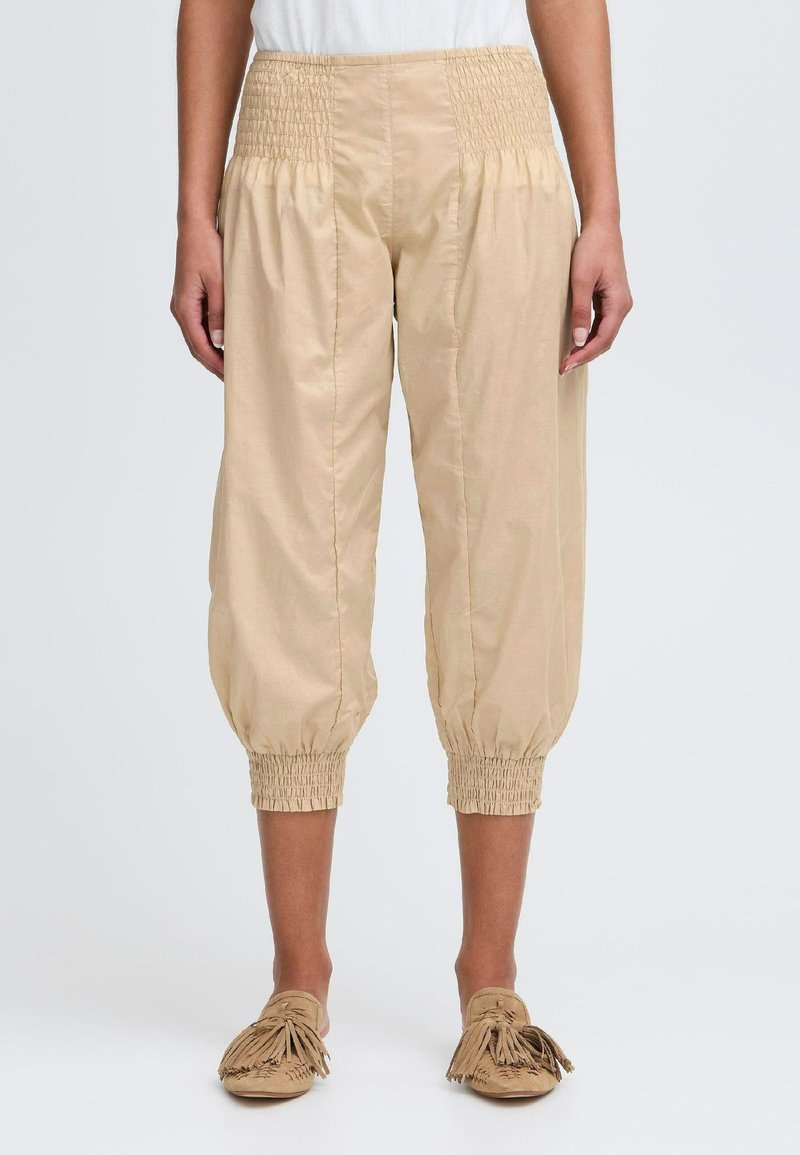 Beige pamut nadrág, smocked derékrésszel és tapered szegélyekkel. Az anyag könnyűnek tűnik, sima textúrával.