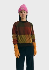 Junge Frau trägt eine rosa Strickmütze, einen mehrfarbigen karierten Pullover, orangefarbene Handschuhe und schwarze Hosen, steht vor einem hellgrauen Hintergrund.