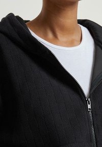 Person trägt einen schwarzen, strukturierten Kapuzenpullover mit Reißverschluss über einem weißen, gerippten Tanktop, wodurch der Hals- und obere Brustbereich sichtbar ist.