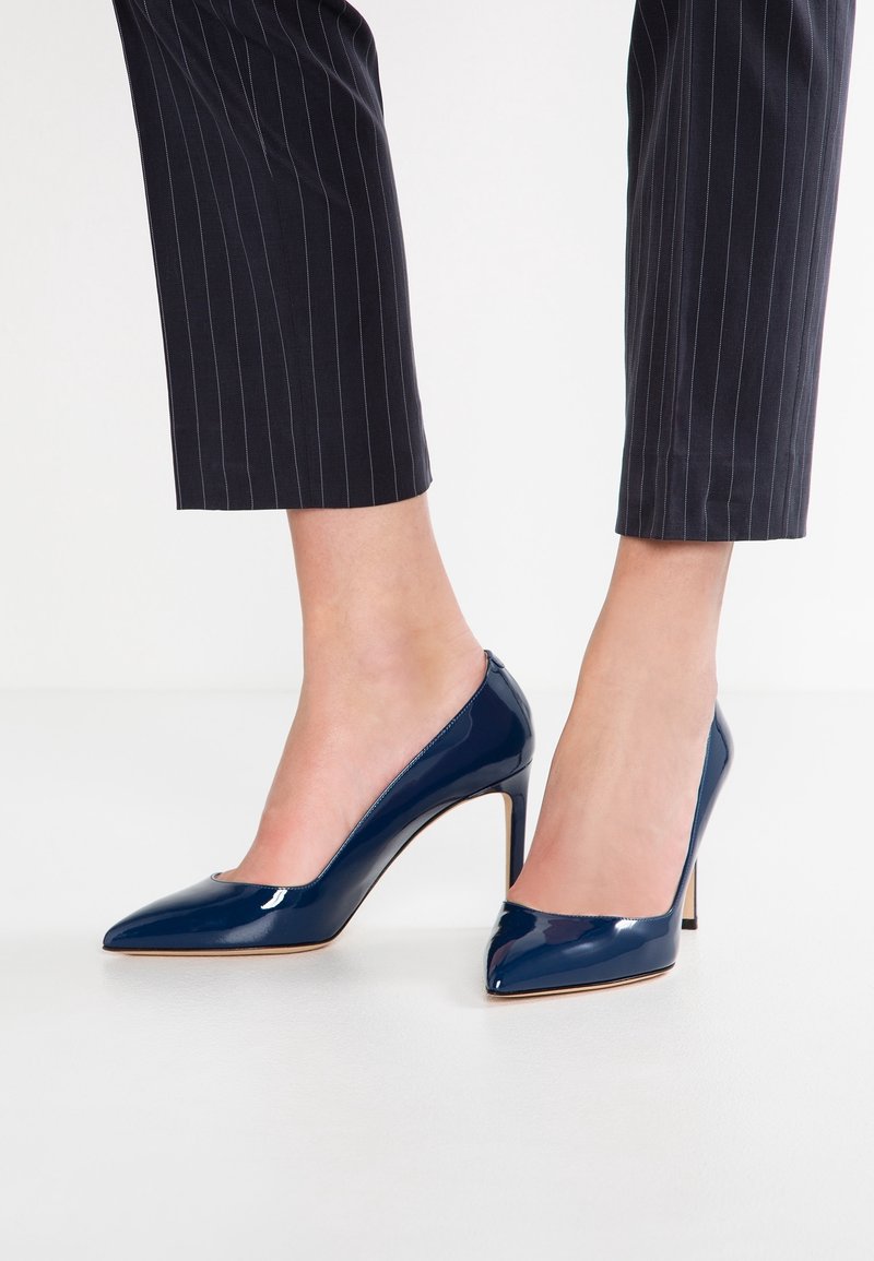 Escarpins en cuir verni bleu marine avec un bout pointu et un talon aiguille fin, posés sur un fond blanc et des pantalons à rayures.