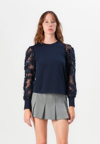 Marineblaues Sweatshirt mit durchsichtigen, floral bestickten Ärmeln, Rundhalsausschnitt und lockerer Passform, kombiniert mit einem grauen, gefalteten Minirock.