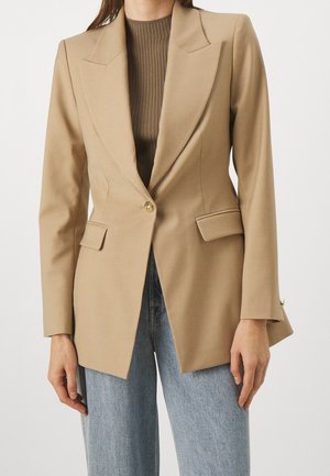Femme portant un blazer beige à un bouton sur un top côtelé marron et un jean bleu clair, montrée du cou aux hanches.