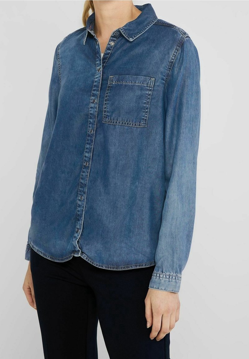 Jeansová košile ve světlé až středně modré barvě, s dlouhými rukávy, s knoflíkovým zapínáním vpředu, s jednou náprsní kapsou, zakřiveným lemem a viditelnými švy.