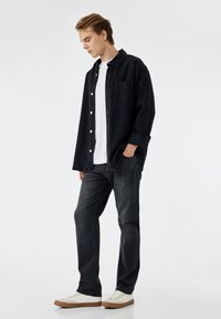 Camisa de mezclilla negra con botones, usada sobre una camiseta blanca, combinada con jeans oscuros y zapatillas blancas con suelas de goma marrón.