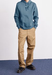 Teal hoodie med framficka och vit logotyp, ihop med beige byxor och svarta snörskor med en texturerad sula.