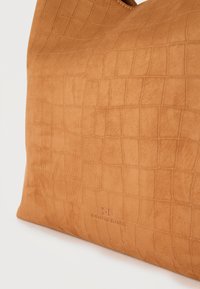 Καφέ ταμπά τσάντα με υφή ψεύτικου κροκοδείλου και διακριτικό ανάγλυφο λογότυπο κοντά στο κάτω κέντρο, σε λευκό φόντο.