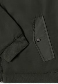 Section d'une veste bomber vert foncé avec un poignet côtelé, une poche plate à fermeture par bouton, et un tissu texturé avec des lignes subtiles.