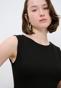 Schwarzes ärmelloses Kleid mit glatter Textur, rundem Ausschnitt und taillierter Silhouette. Das Model trägt goldene Creolen.