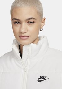 Biała ocieplana kurtka z wysokim kołnierzem, zapięciem na zamek błyskawiczny i czarnym logo Nike na lewej piersi. Gładka tekstura i nowoczesny design.