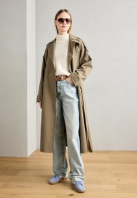 Khaki trenchcoat, kremfarget ribbestrikket genser, lyseblå jeans og blå sneakers. Store solbriller og et brunt belte fullfører stilen.