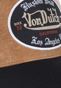 Tissu en feutre marron avec un sommet en tissu noir, présentant un logo circulaire Von Dutch en crème et rouge, ainsi qu'un texte détaillé en dessous.