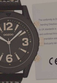 Montre en silicone noire avec de grands chiffres blancs, trois boutons-poussoirs en ton or, et un bracelet texturé noir. Comprend une documentation conforme aux normes CE.