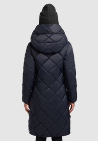Inimene, kellel on seljas pikk, vatitatud tumesinine mantel kapuutsiga ja tume kudumismüts, on nähtav seljaga lihtsa taustaga.