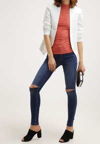 Blazer bianco aderente, top coral texturizzato, jeans skinny blu strappati, mules a tacco blocco neri, con una piccola pochette nera.