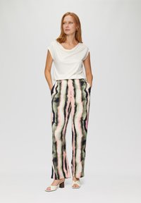 Haut blanc à manches courtes avec encolure drapée ; pantalon large à motif présentant des rayures verticales roses, vertes et noires ; le mannequin pose les mains dans les poches.