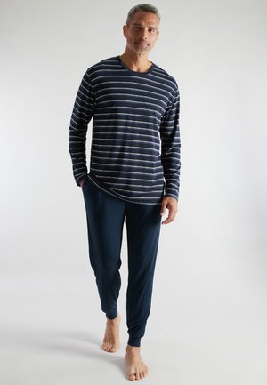 Mann steht barfuß und trägt ein marineblau gestreiftes Langarmshirt und marineblaue Joggerhosen vor einem einfarbigen hellen Hintergrund.