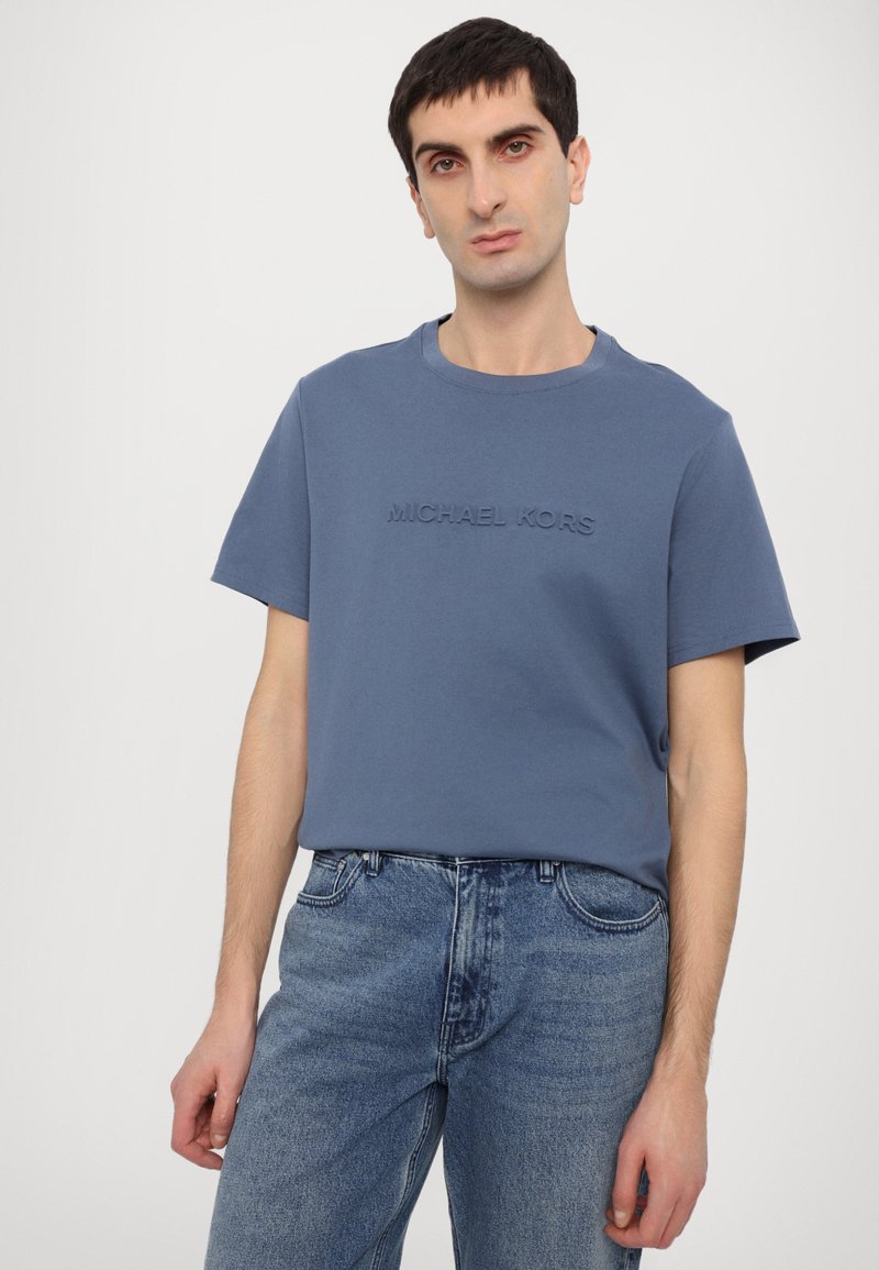 Blaues Baumwoll-T-Shirt mit Rundhalsausschnitt, auf der Vorderseite mit dem geprägten Text "MICHAEL KORS". Kombiniert mit hellen Jeans.