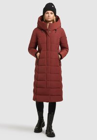 Vrouw in een lange kastanjebruine gewatteerde winterjas met capuchon, zwarte beanie, zwarte leggings en zwarte chunky laarzen, staande tegen een witte achtergrond.