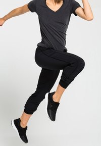 Un t-shirt noir à col en V associé à un pantalon de jogging noir avec des poignets élastiques et une texture lisse. Des chaussures de sport noires complètent la tenue.