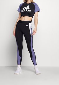 Czarny crop top z białym logo Adidas, w połączeniu z czarnymi legginsami z fioletowymi panelami bocznymi i dopasowanymi akcentami. Materiał wydaje się elastyczny.