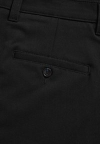 Gros plan sur un tissu noir montrant une poche arrière de pantalon avec une fermeture par bouton et une couture de passant de ceinture en haut.