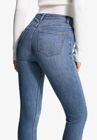 Jean en denim bleu taille haute, coupe ajustée, avec poches arrière, coutures décoratives et texture lisse.