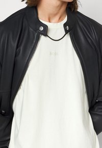 Czarna skórzana kurtka typu bomber z wysokim kołnierzem i srebrnym zamkiem błyskawicznym, noszona na jasnobeżowej koszuli z subtelnym wytłoczonym logo „BOSS”.