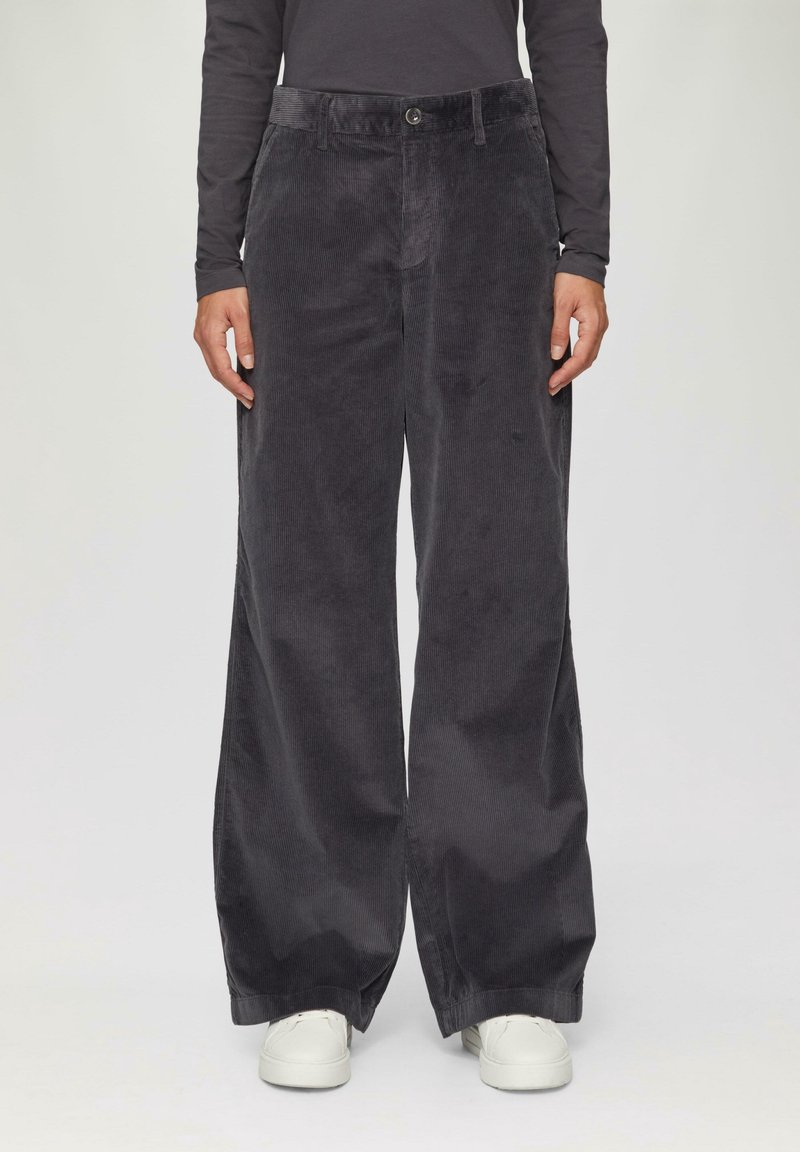 Personne portant un pantalon large en velours côtelé gris foncé, un haut noir à manches longues et des baskets blanches, debout devant un fond uni.