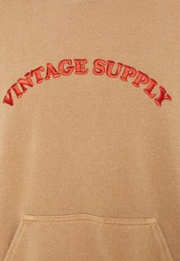 Brown pullover hoodie with a soft texture, featuring red curved "VINTAGE SUPPLY" embroidery and a kangaroo pocket at the front.