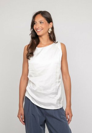 Femme souriante aux longs cheveux bruns portant des boucles d'oreilles en or, un haut sans manches en soie blanche et un pantalon gris ample sur un fond clair uni.