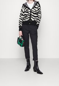 Černobílý pletený kardigan se vzorem zebra s kapucí, v kombinaci s úzkými černými kalhotami a černými kotníkovými botami na podpatku. Doplňuje ho zeleno-černá kabelka.