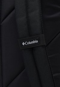 Černý prošívaný batoh Columbia s texturovanou tkaninou, polstrovanými popruhy a bílou visačkou s logem. Odolný a funkční design.