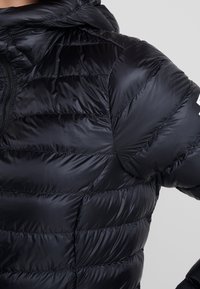 Svart pufferjacka med horisontella quiltade sektioner, luva och dragkedja fram. Materialet är slätt och glansigt och har ett logotypaccent.