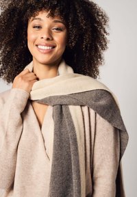 Sciarpa a maglia con texture a coste, con strisce di crema, grigio e grigio scuro. Indossata in modo informale sopra un maglione beige chiaro.