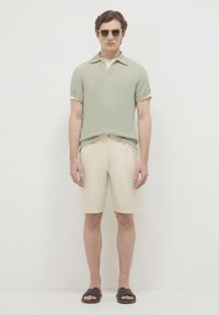 Polo shirt verde chiaro a maniche corte, pantaloni corti beige e sandali scivolati marrone scuro. Design minimalista con colletto ripiegato e texture morbida.