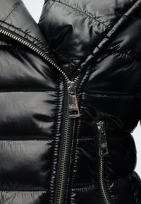 Veste noire matelassée avec deux curseurs de fermeture éclair DKNY argentés, l’un partiellement fermé et l’autre complètement fermé sur une poche visible.